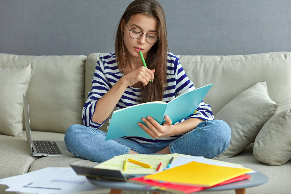 Brunette teenage girl with glasses studying on sofa, writing in workbook with pen, computer beside her and desk with books in front.