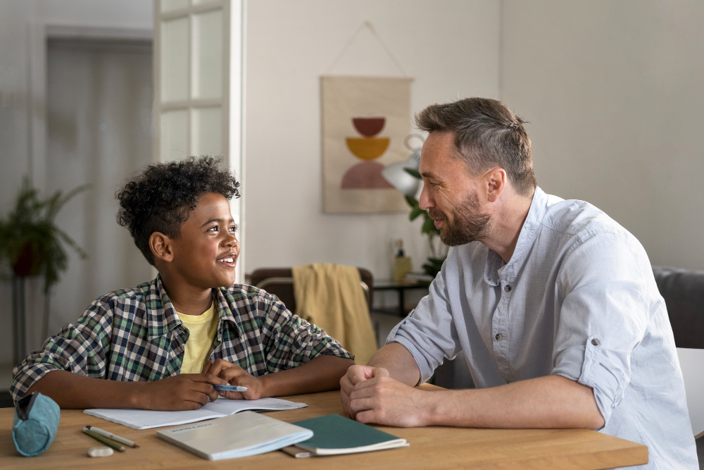 Smiling father and child sitting together at a desk at home. The child is holding a pen and has a school workbook open in front of them, appearing engaged in learning.