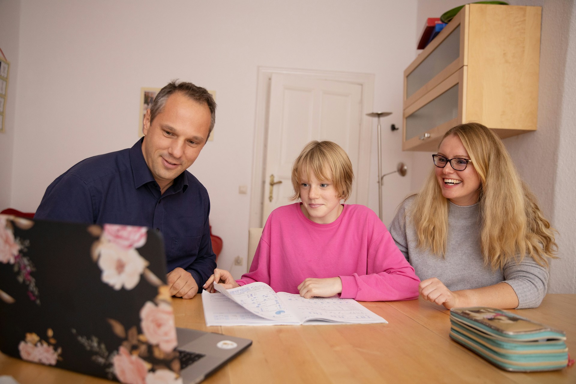 Parents helping teenage daughter with homework on laptop at home, smiling together at desk.