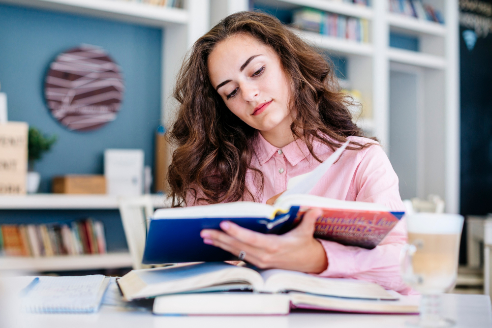 Teenage girl in a pink shirt studying at a library desk, reading a book with two other open books stacked underneath.